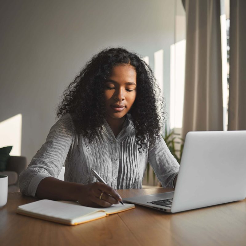 beautiful-mixed-race-student-girl-with-voluminous-hairdo-working-home-assignment-her-room-sitting-wooden-desk-using-laptop-writing-down-copybook-people-technology-education